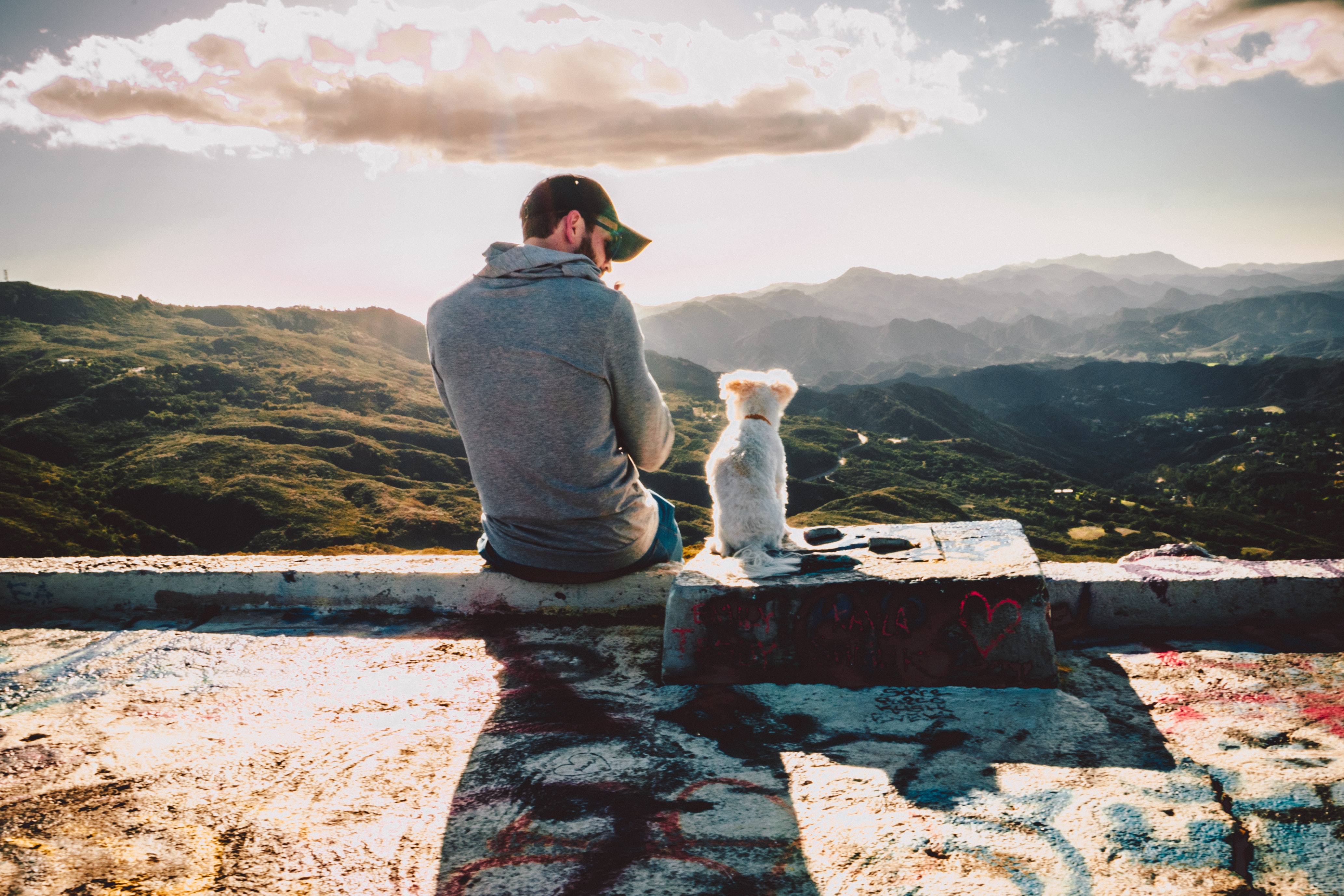 Man and his dog sitting on a ledge overlooking a scenary of mountains. Man passionately looks at his dog.